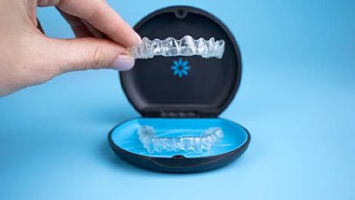 Two fingers grasping an Invisalign tray from its case with light blue background